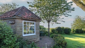 A stone cottage with a tiled roof, a large window, and a garden with a tree and bushes.