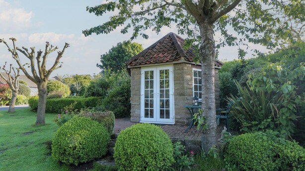 A small stone building with a tiled roof, surrounded by well-maintained hedges and trees.