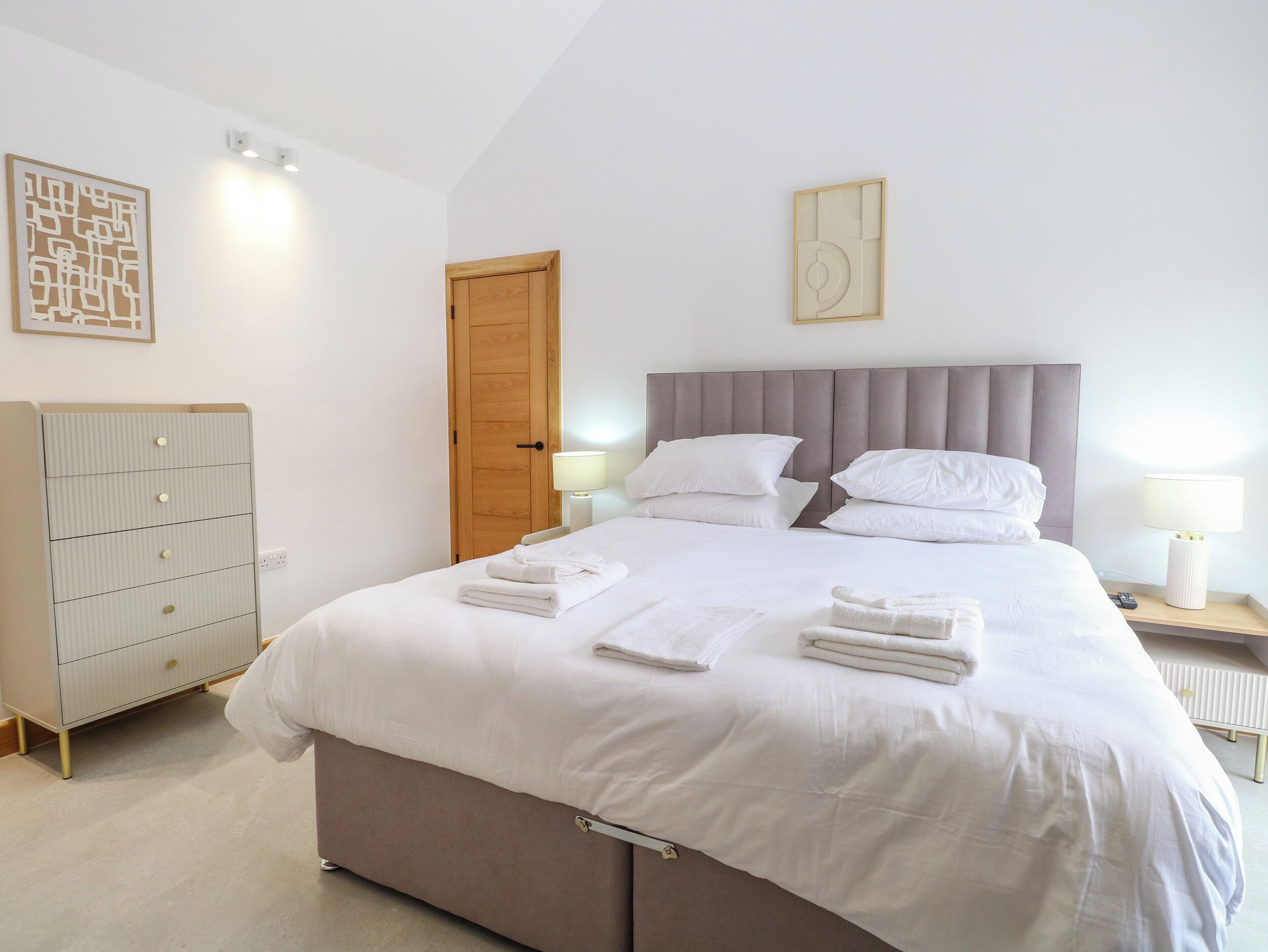 A neatly made bed with white linens and towels, a wooden wardrobe, and a door in a simple, clean room.
