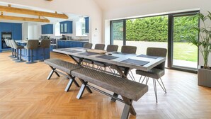 A modern kitchen with blue cabinets and a large island, a dining area with a long table and benches, and a view of a green landscape through large windows. - County Farm Barn (Norwich)