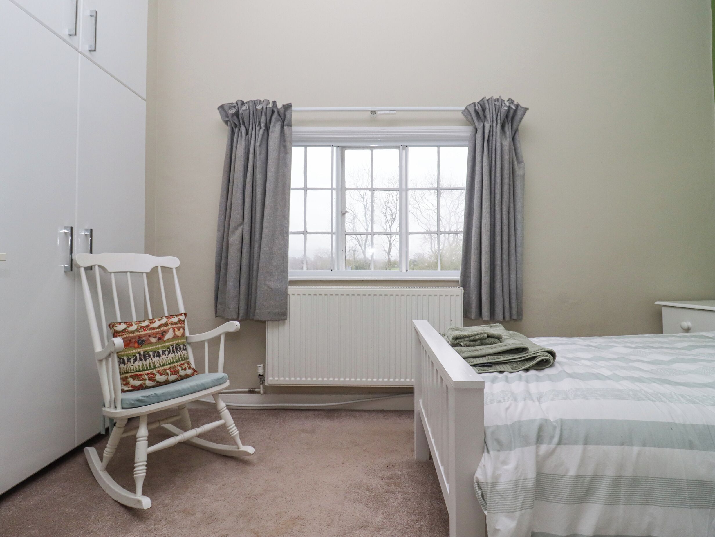 A bedroom with a white wardrobe, a rocking chair, a window with curtains, a radiator, and a bed with a striped bedspread.
