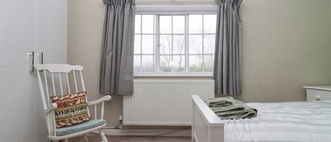 A bedroom with a white wardrobe, a rocking chair, a window with curtains, a radiator, and a bed with a striped bedspread.