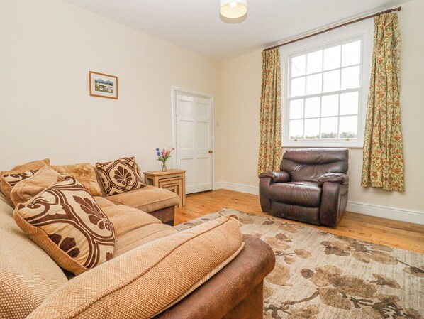 A living room with a sofa set, a leather armchair, a small side table, and a window with floral curtains. - The Shires at Bickerscourt (Solihull)