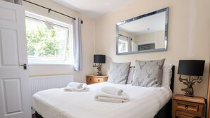 Un lit bien fait, avec du linge de lit blanc et des serviettes pliées, une table de chevet avec une lampe, et une fenêtre donnant sur un paysage verdoyant.