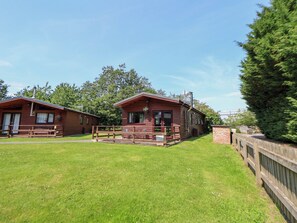 A wooden cabin with a porch and a small deck, surrounded by a wooden fence and a grassy lawn. - The Fallows (Saint Asaph)