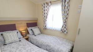 Une chambre avec deux lits simples, une table de chevet avec une lampe, une literie à motifs, une fenêtre avec des rideaux et une tête de lit en bois.