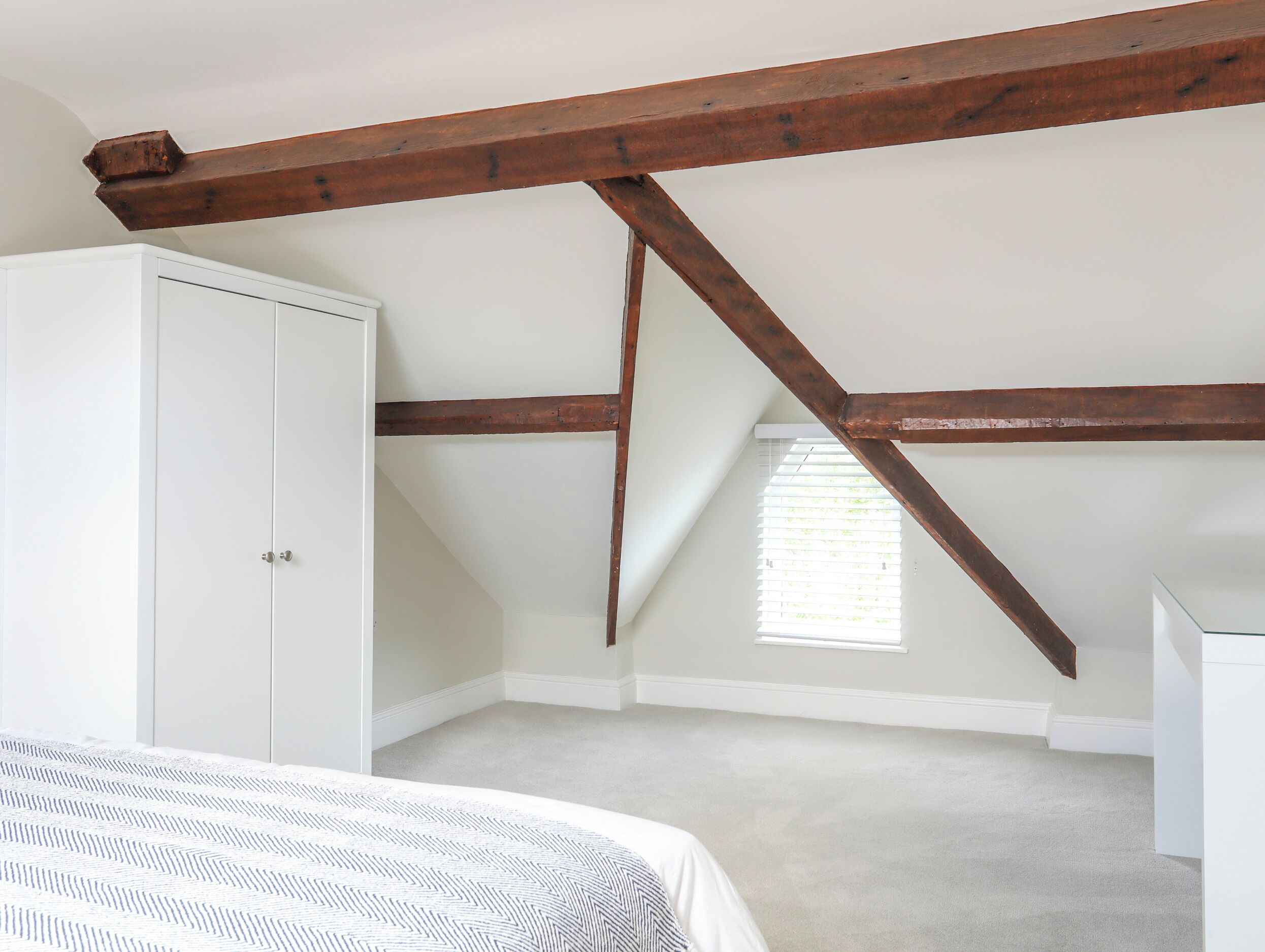 Een slaapkamer met een schuine kap, een garderobe, een bed met een bedovertrek met patroon, en een raam met jaloezieën.