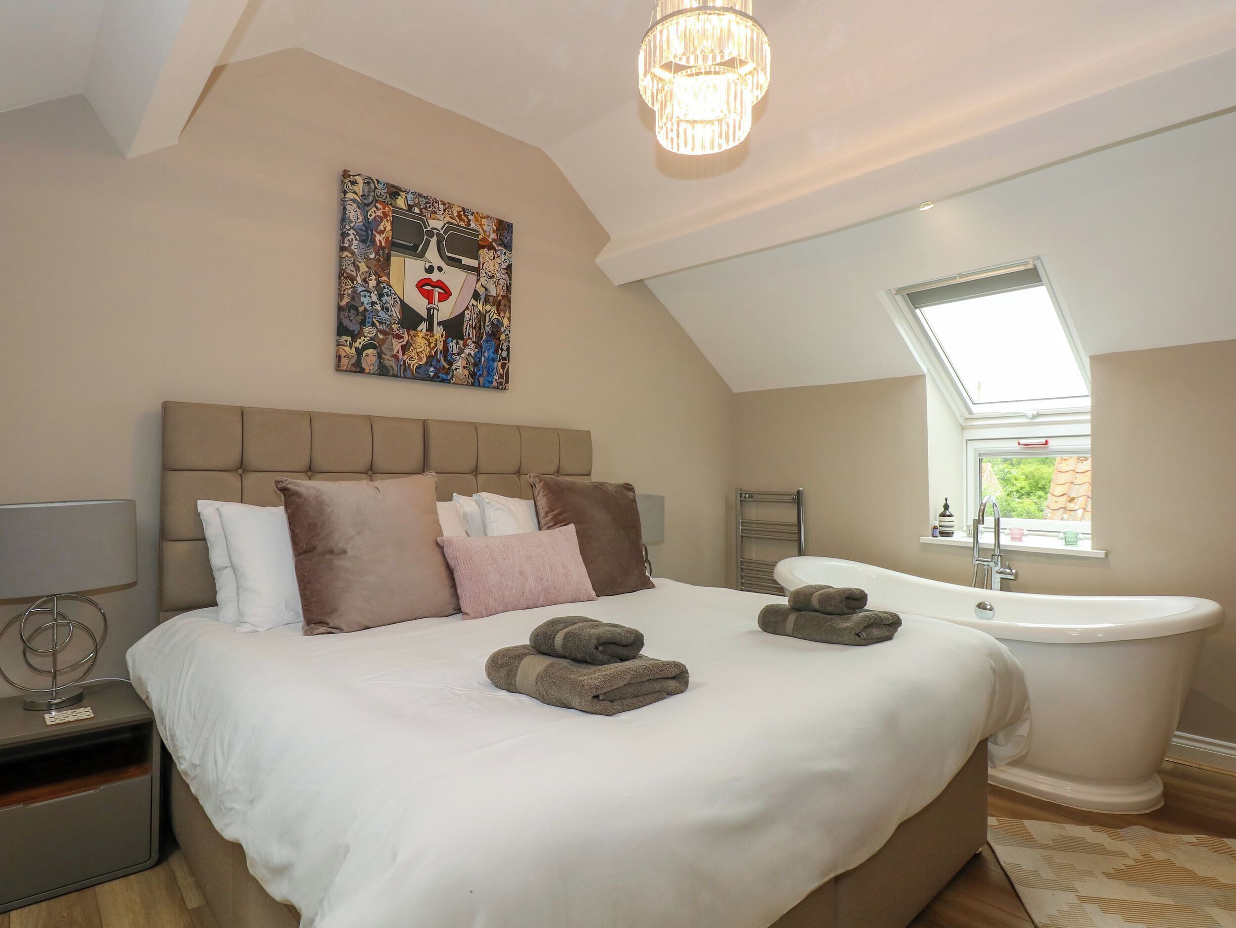 Una camera da letto con un letto grande, un comodino, una lampada, un quadro, un lampadario, una finestra e un bagno con una vasca.