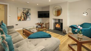 A modern living room with a sofa, a glass coffee table, a TV, a fireplace, and a teal armchair. - Daisy's Place (Fakenham)