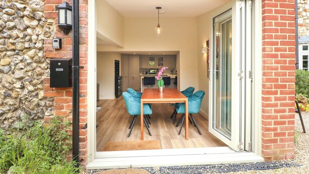 A dining area with a table and chairs, a kitchen in the background, and a stone wall. - Daisy's Place (Fakenham)