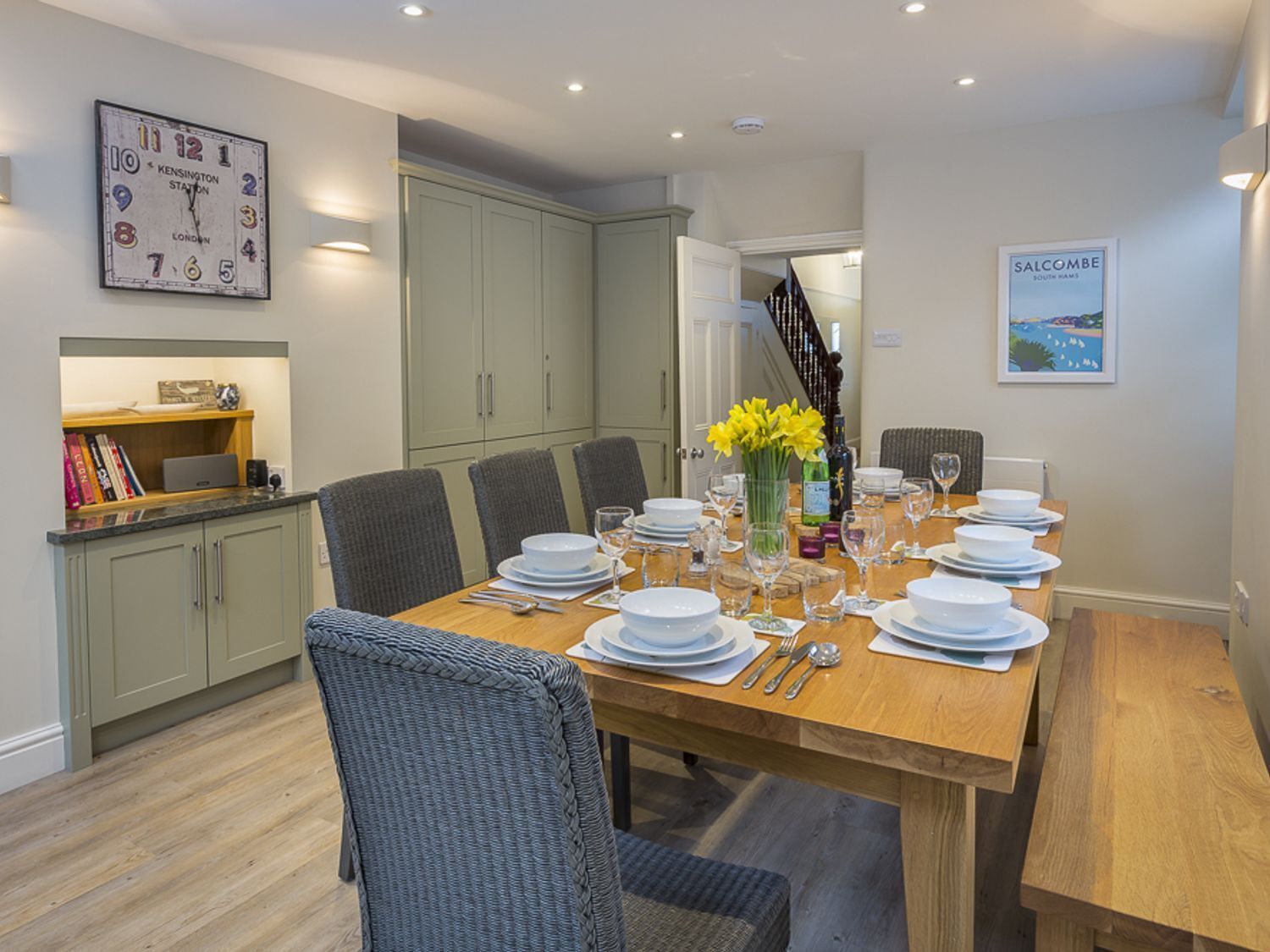 A dining area with a wooden table set for six, surrounded by grey chairs. A wall clock and a framed picture are visible.