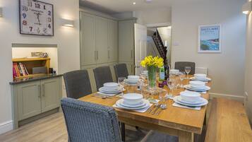 A dining area with a wooden table set for six, surrounded by grey chairs. A wall clock and a framed picture are visible.