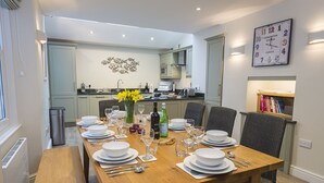 A dining room with a wooden table set for four, surrounded by chairs. A kitchen is visible in the background with modern appliances and a decorative wall piece. - Leylands (Salcombe)