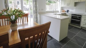 A modern kitchen with a central island, white cabinetry, and a dining area with wooden chairs and a vase of flowers. - Cottage on the Green (Caernarfon)
