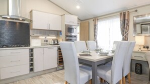 A modern kitchen with white cabinets, a built-in oven, and a microwave. There is a dining table with four chairs and a chandelier. - Tawny Owl Lodge (Windermere)