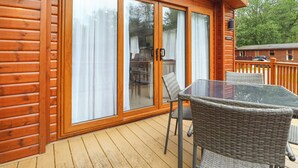 A wooden cabin with a glass door, a deck with wicker furniture, and a clear view of the surrounding trees. - Tawny Owl Lodge (Windermere)