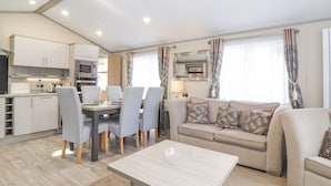 A modern living room with a sofa, dining table, and chairs, a kitchen area with appliances, and a large mirror. - Tawny Owl Lodge (Windermere)