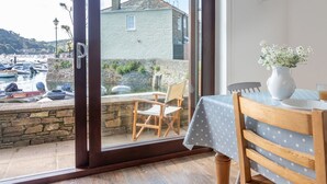 Una habitación con puerta corrediza de vidrio que da a un patio con mesa y sillas, un florero con flores y vista a un puerto con barcos y edificios.