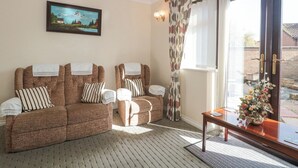 A room with two brown sofas, a wooden table with a vase, a painting on the wall, and a window with curtains. - Fatima (Burnham-on-Sea)