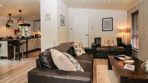 A modern living room with a brown leather sofa, a wooden coffee table, and a black leather couch. There is a kitchen area with a bar stool and a window with blinds. - Somerford Lodge (Windermere)