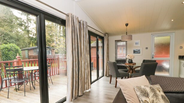 A spacious living area with a large sliding glass door leading to a deck, a dining area with a table and chairs, and a cozy seating area with a sofa and cushions. - Somerford Lodge (Windermere)