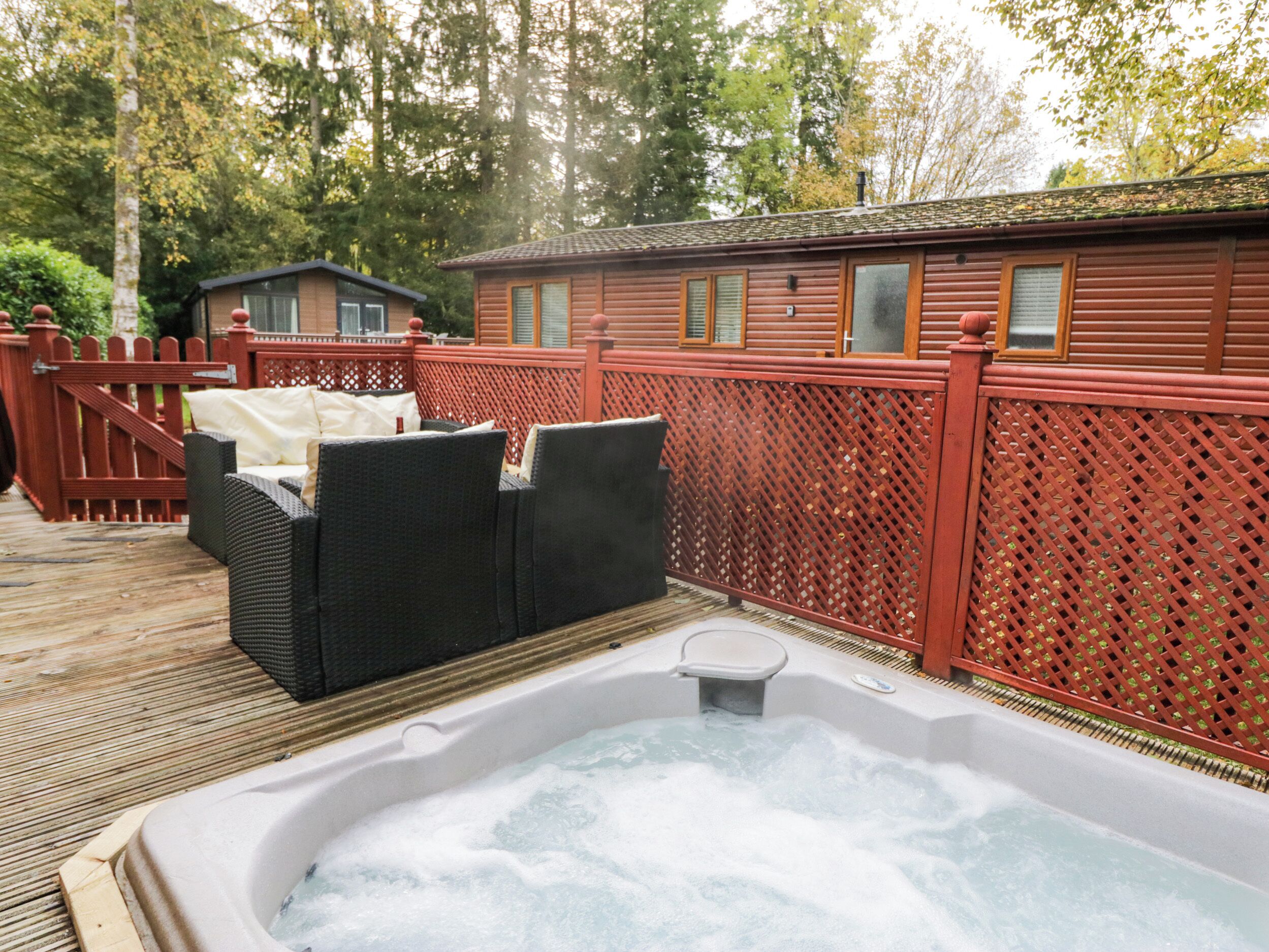 A hot tub on a wooden deck with wicker furniture, surrounded by a red fence and a small building in the background.