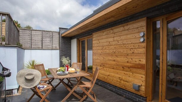 Une terrasse avec une table et des chaises en bois, un barbecue et un sol en pierre.