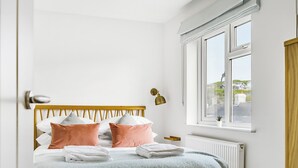 A neatly made bed with a wooden headboard, a window with a view of buildings, and a radiator below. - Fairwinds (Redruth)