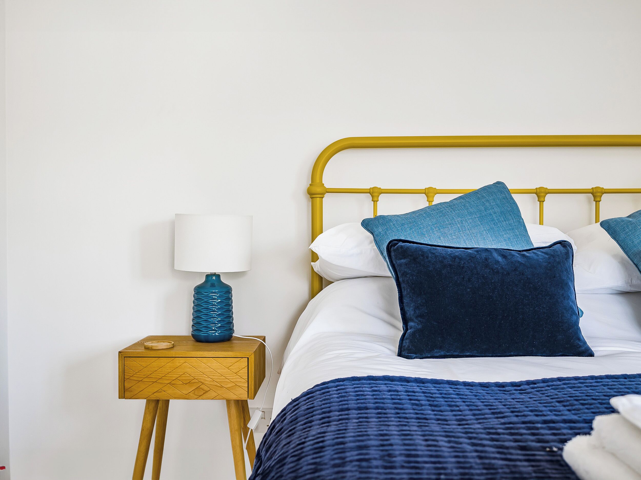 Ein Bett mit gelbem Metall-Kopfstück, weißer Bettwäsche und blauen Kissen. Am Bett steht ein Nachttisch aus Holz mit einer blauen Lampe.
