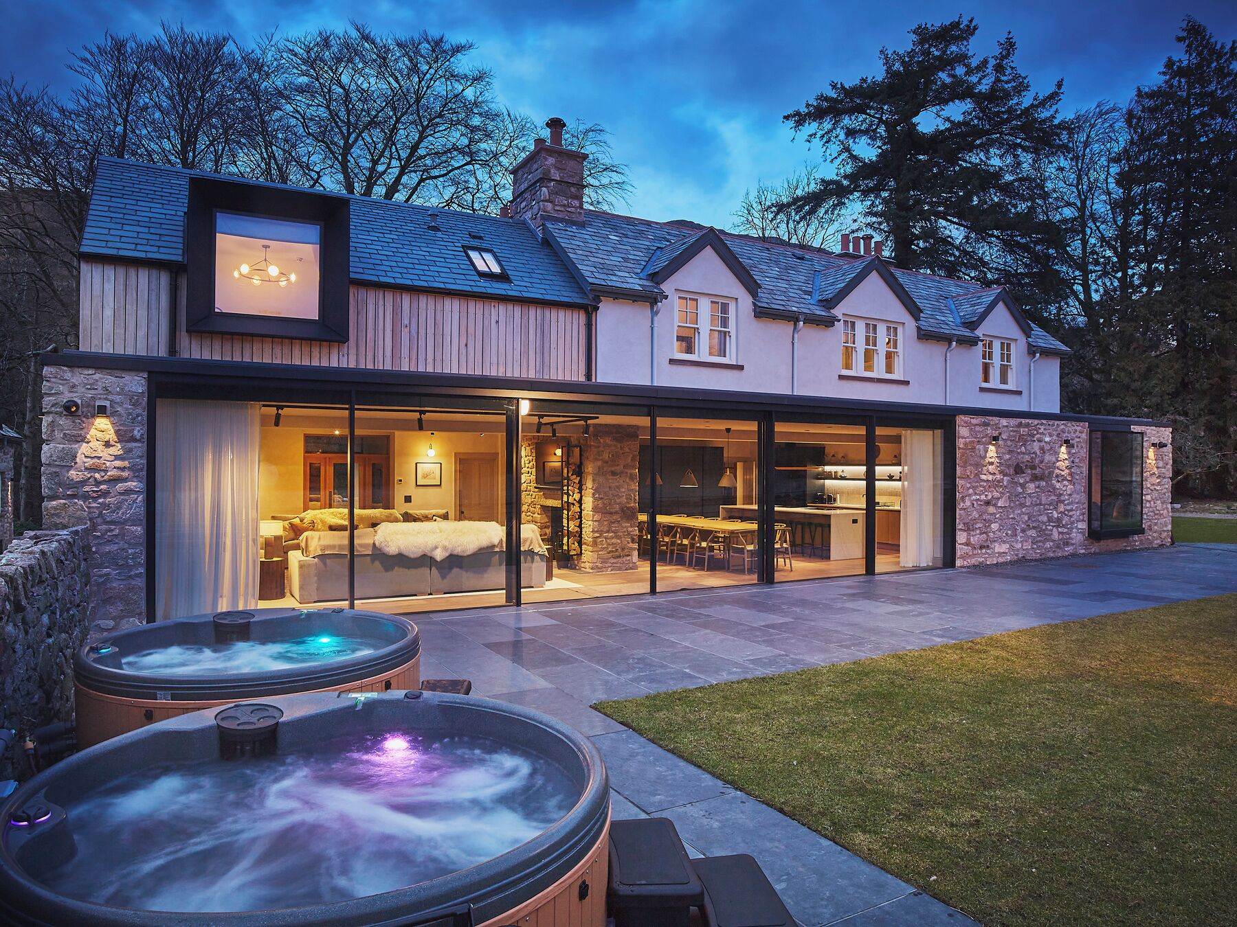 A modern house with a large glass door leading to a dining area and a bedroom. There are two hot tubs in the foreground.