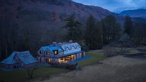 A large, modern house with a stone facade and multiple chimneys, surrounded by a fenced property and a mountainous backdrop. - Eskdale Retreat (Holmrook)