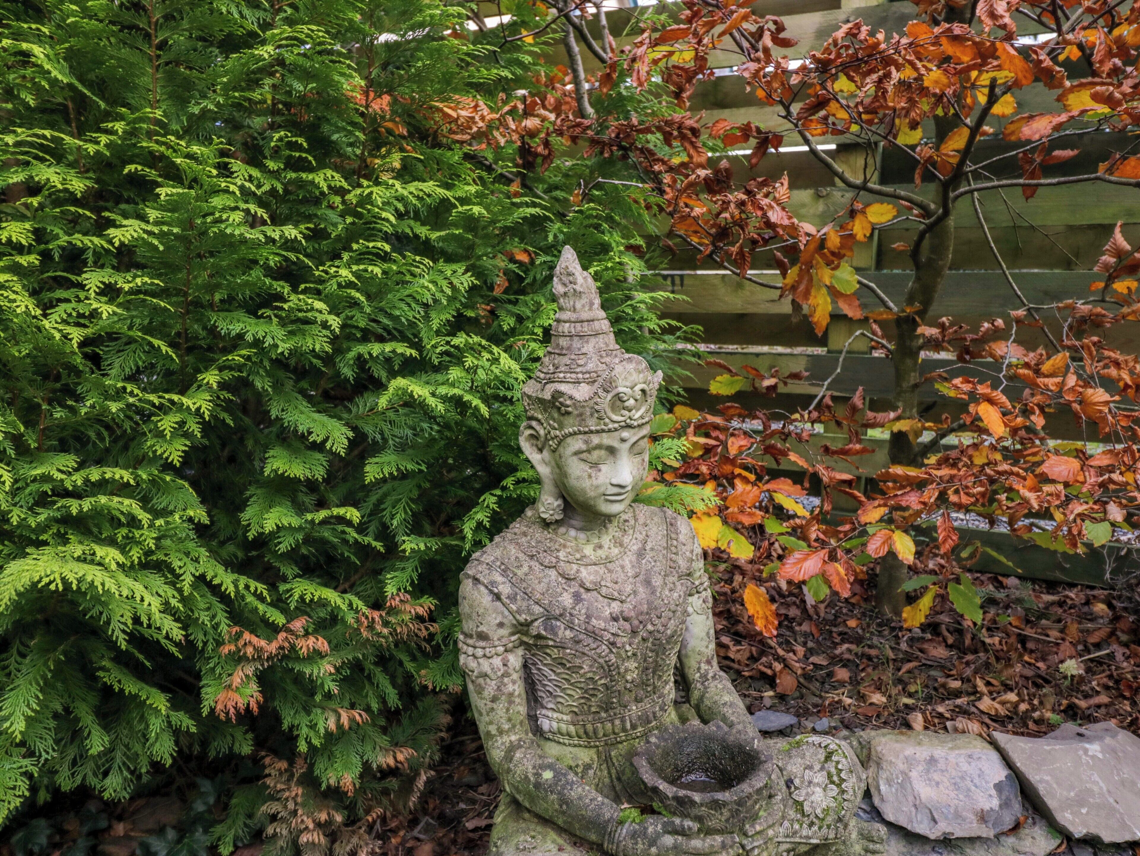 A stone statue of a seated figure with a bowl in hand, surrounded by lush greenery and autumn leaves.