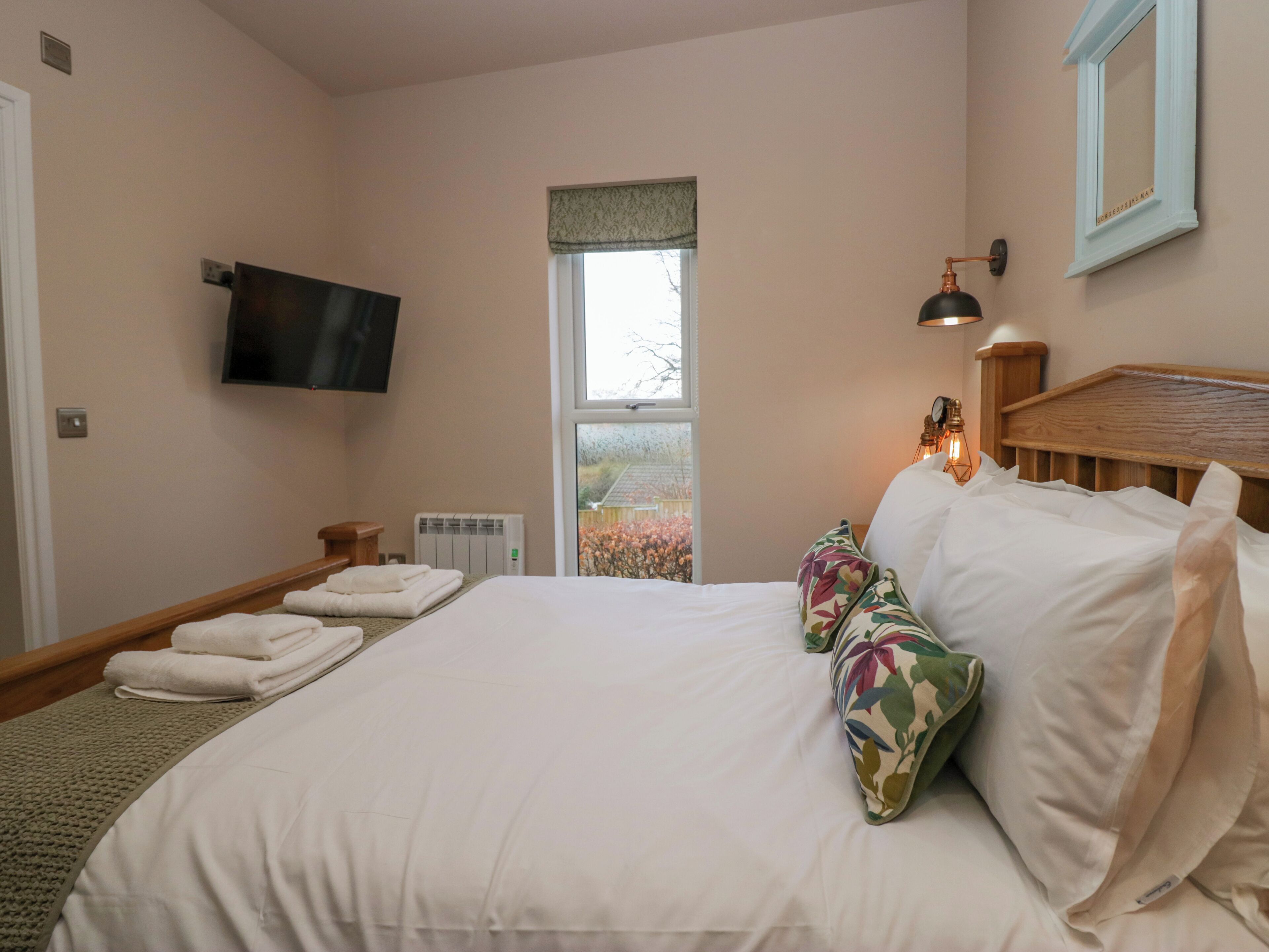 A hotel room with a bed, a flat-screen TV mounted on the wall, a window with a view of trees, and a bedside lamp.