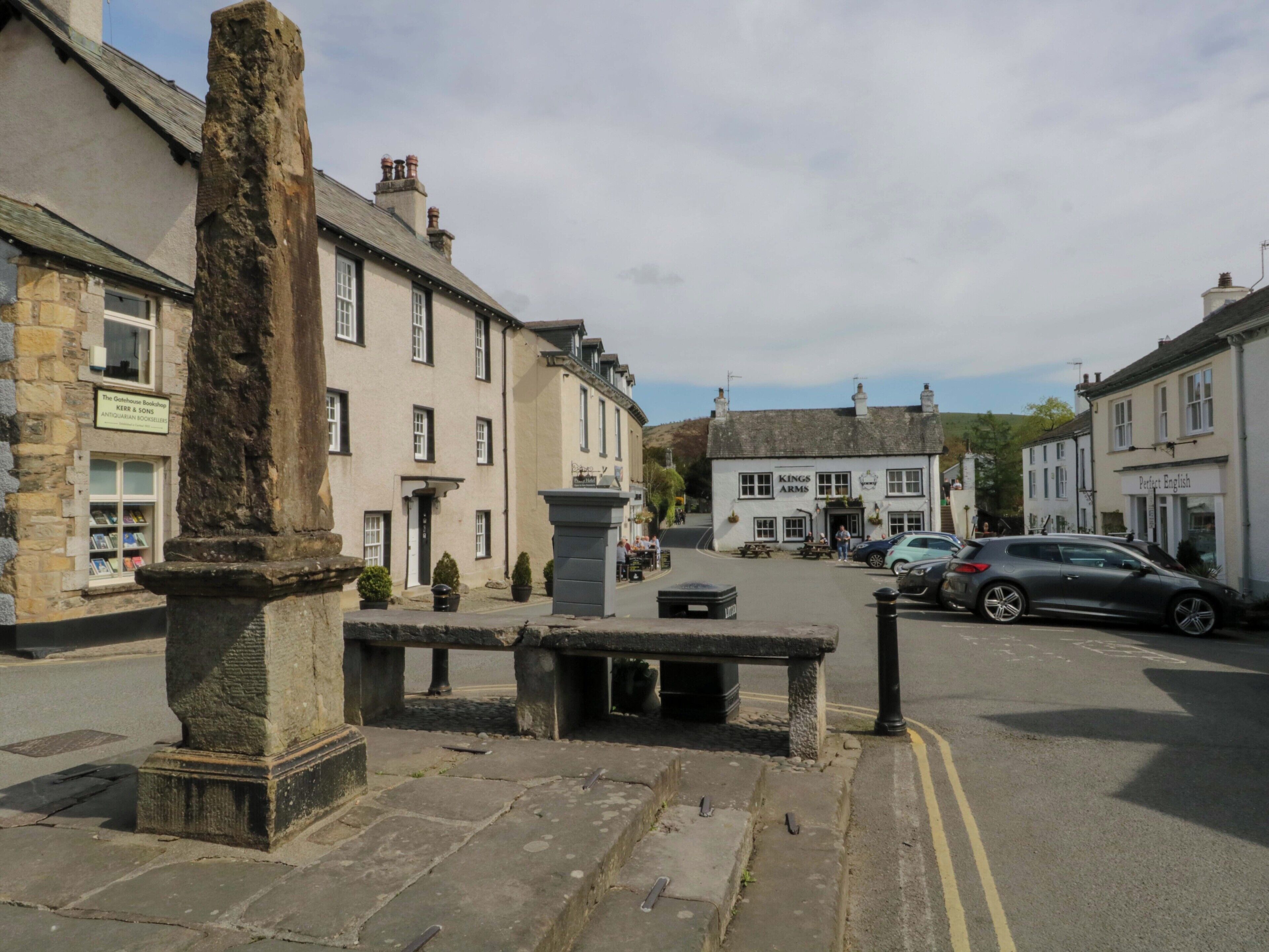 Une rue avec un monument en pierre, un bâtiment blanc portant l’inscription « Kings Arms » et doté d’une terrasse, et une enseigne indiquant « Perfect English ».