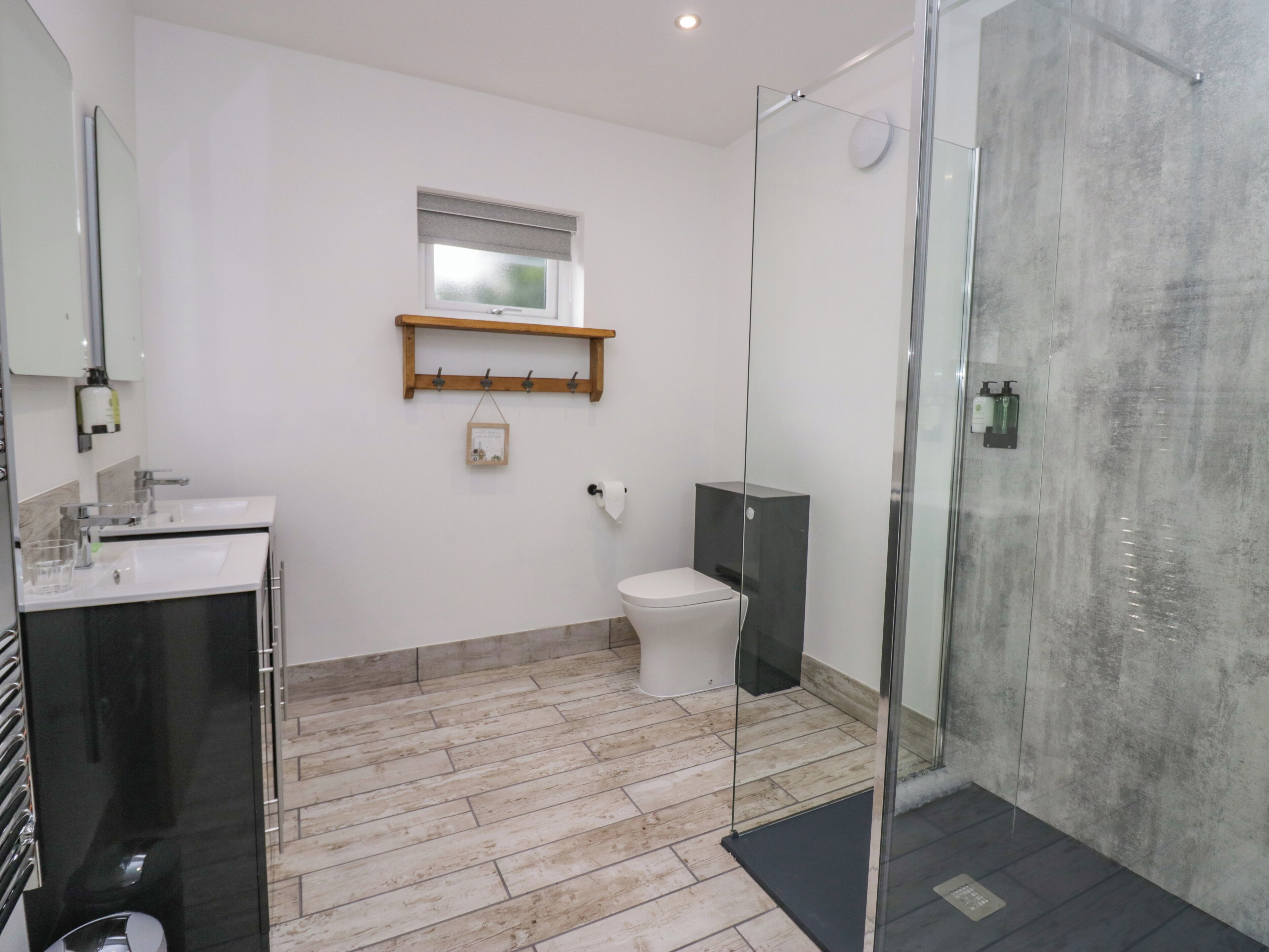 A modern bathroom with a glass shower enclosure, a white toilet, and a wooden shelf above the sink.