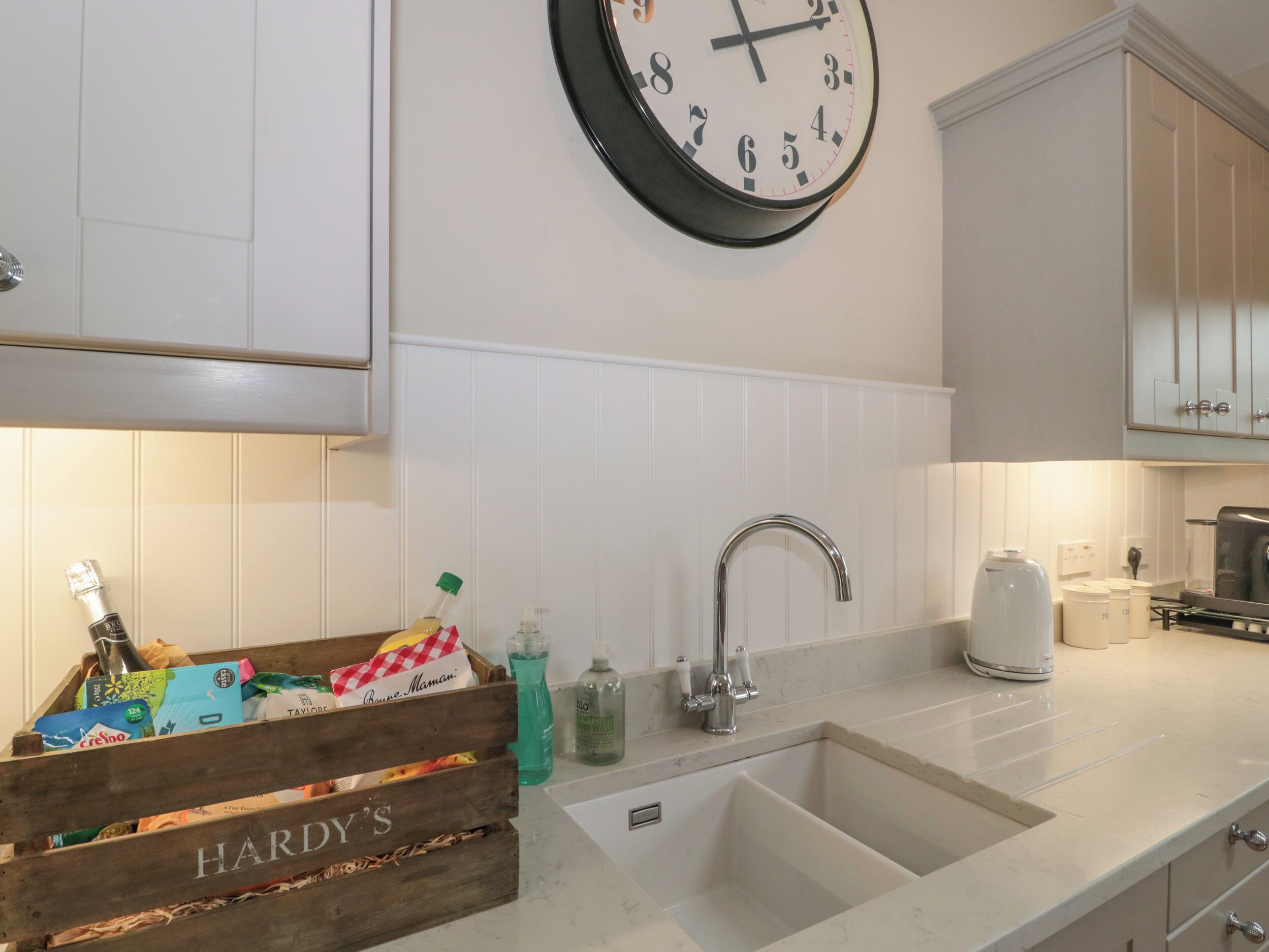 Une cuisine avec des armoires blanches, un évier à deux bacs, un robinet en acier inoxydable, une horloge et une caisse en bois portant l’inscription « HARDY’S » et contenant divers objets.