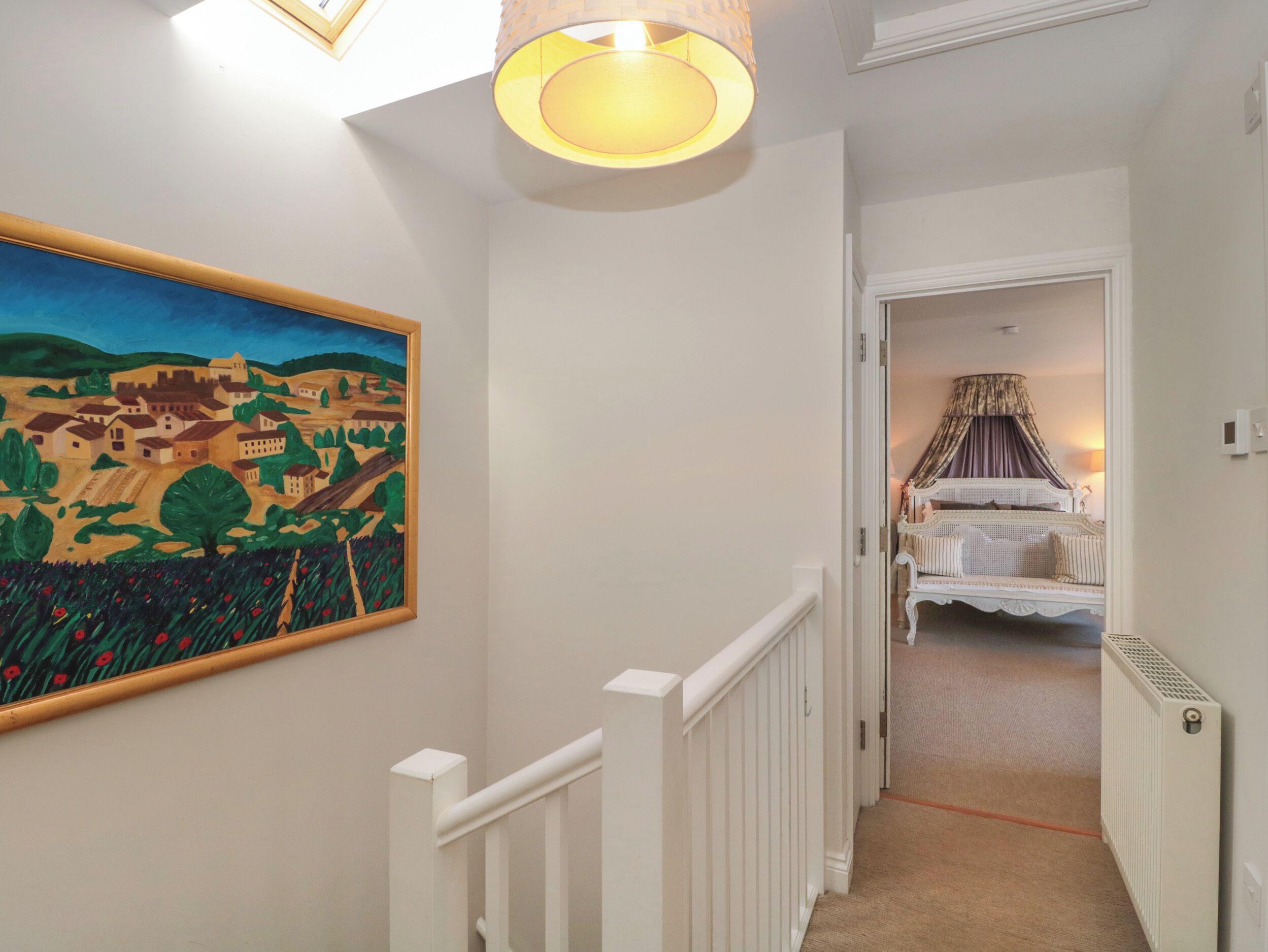A hallway with a painting of a landscape, a staircase, and a bedroom with a bed and curtains.