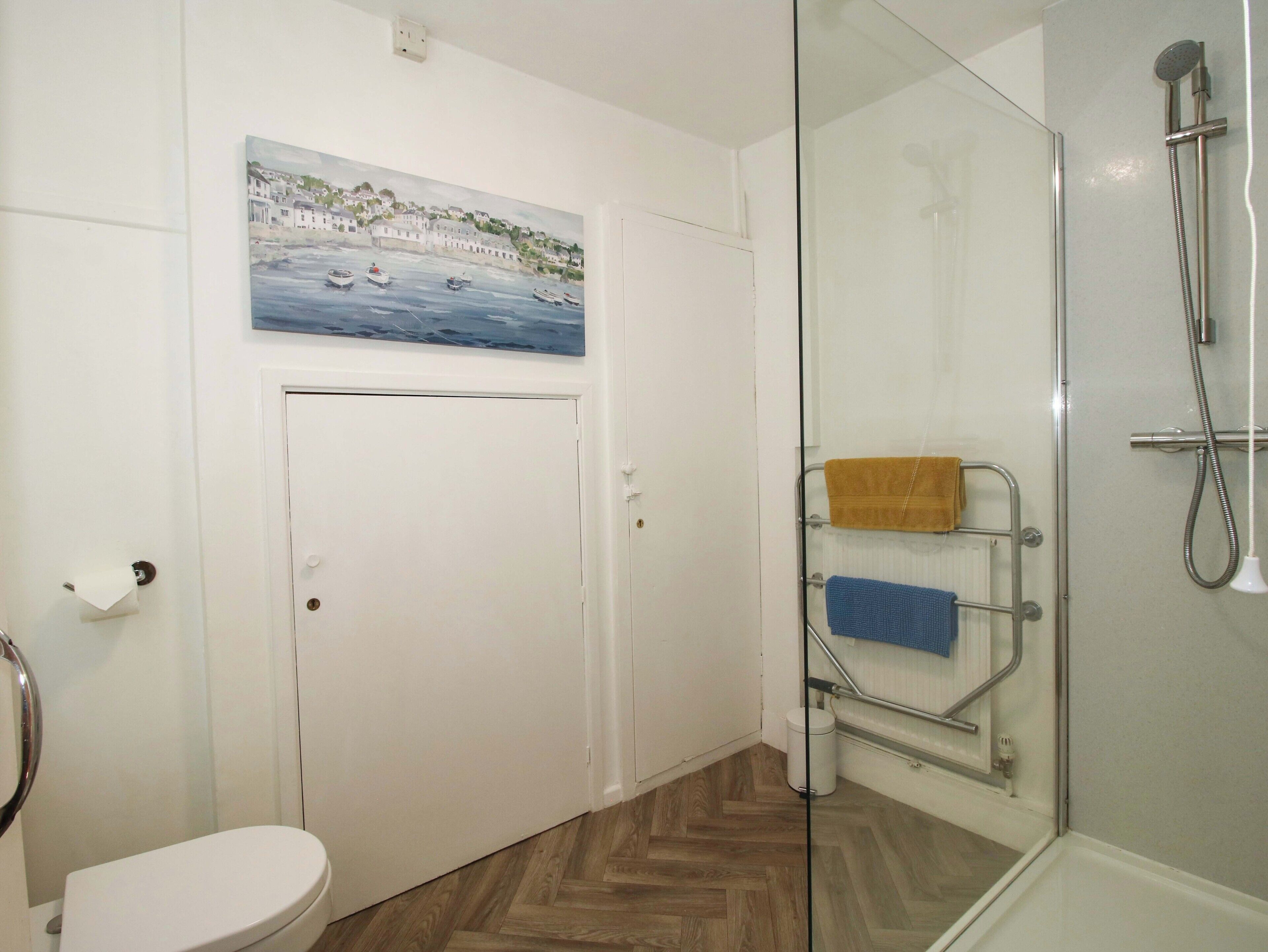 A bathroom with a glass shower enclosure, a toilet, and a painting of a coastal scene.