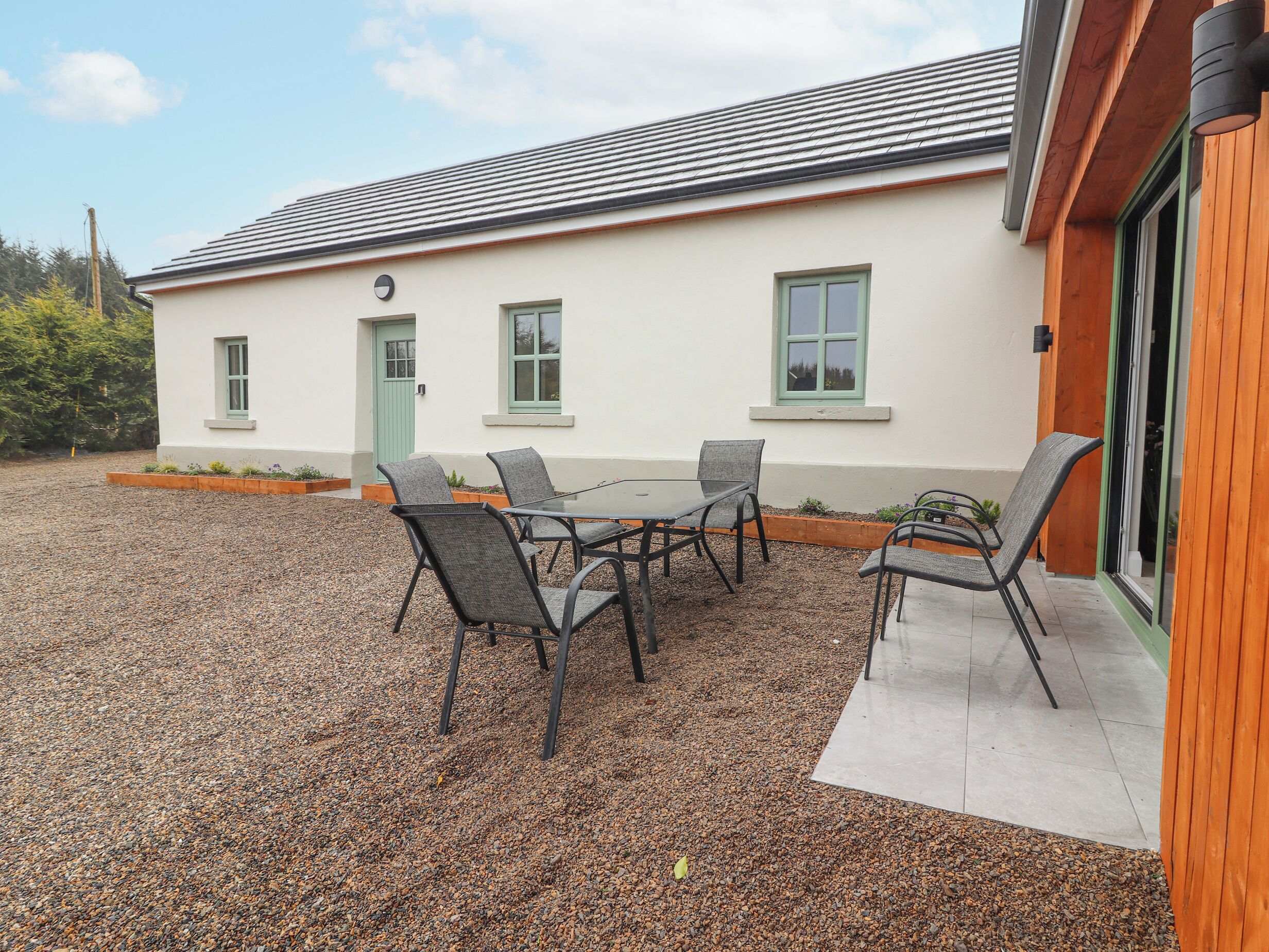 A building with a white exterior, a green door, and a window with a green frame. There is a gravel patio with a table and chairs.