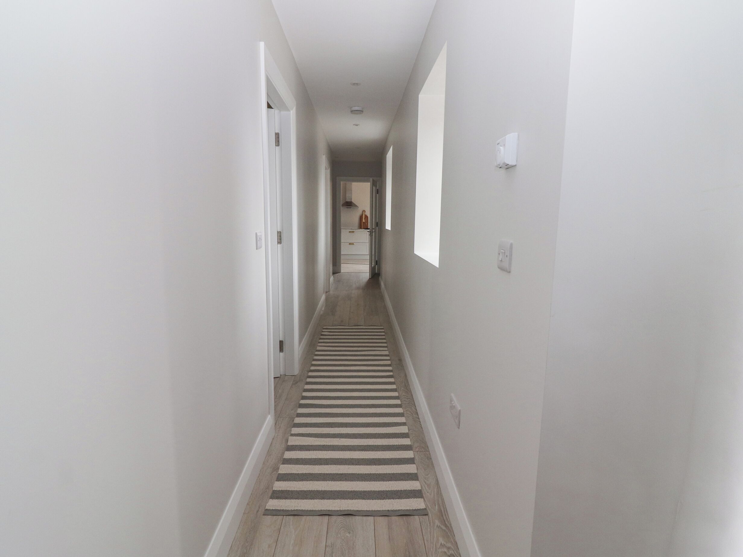 A narrow hallway with a striped rug, white walls, and a door leading to a room.