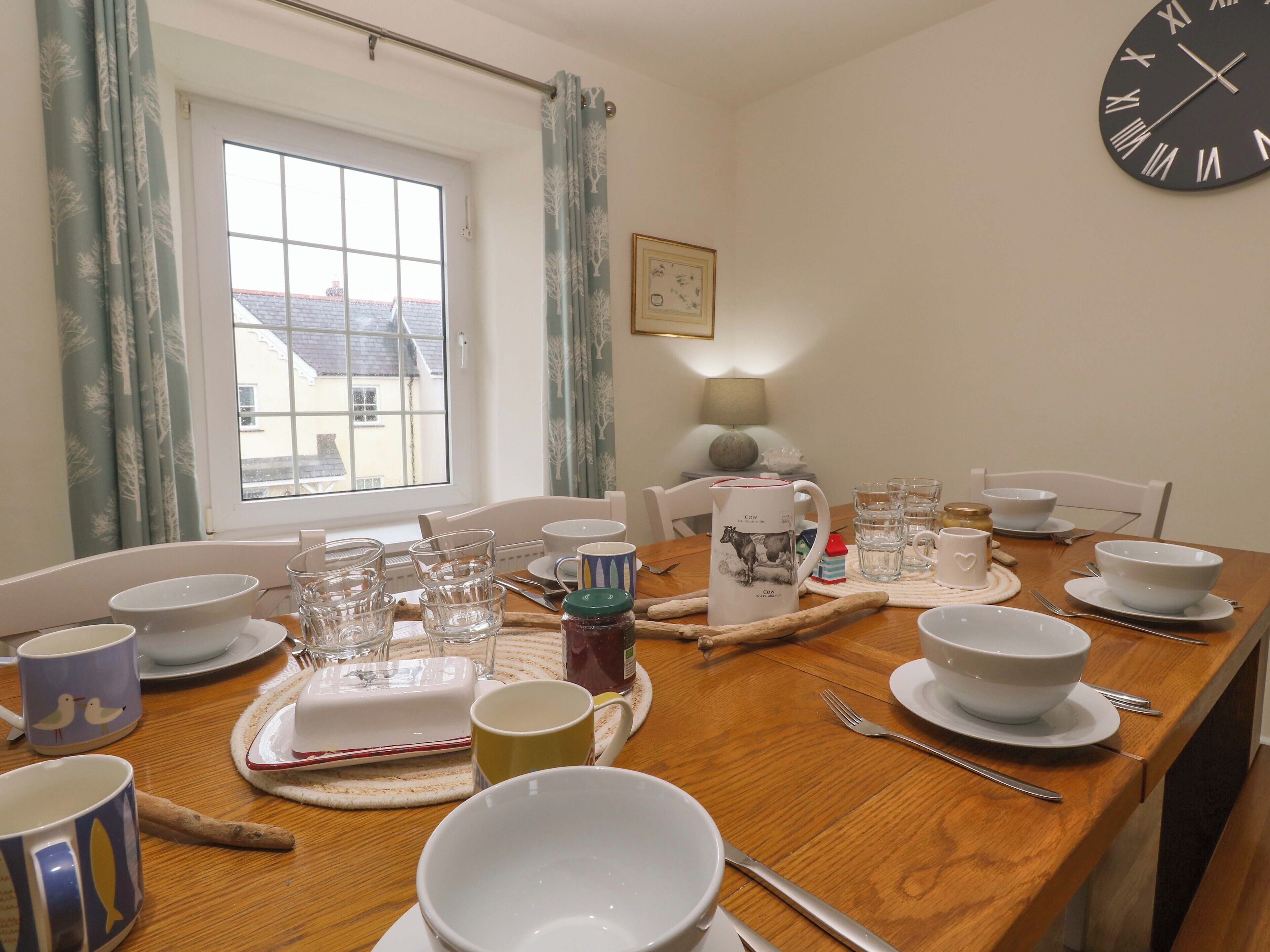 A dining table set with bowls, cups, and cutlery, a clock on the wall, and a window with curtains.