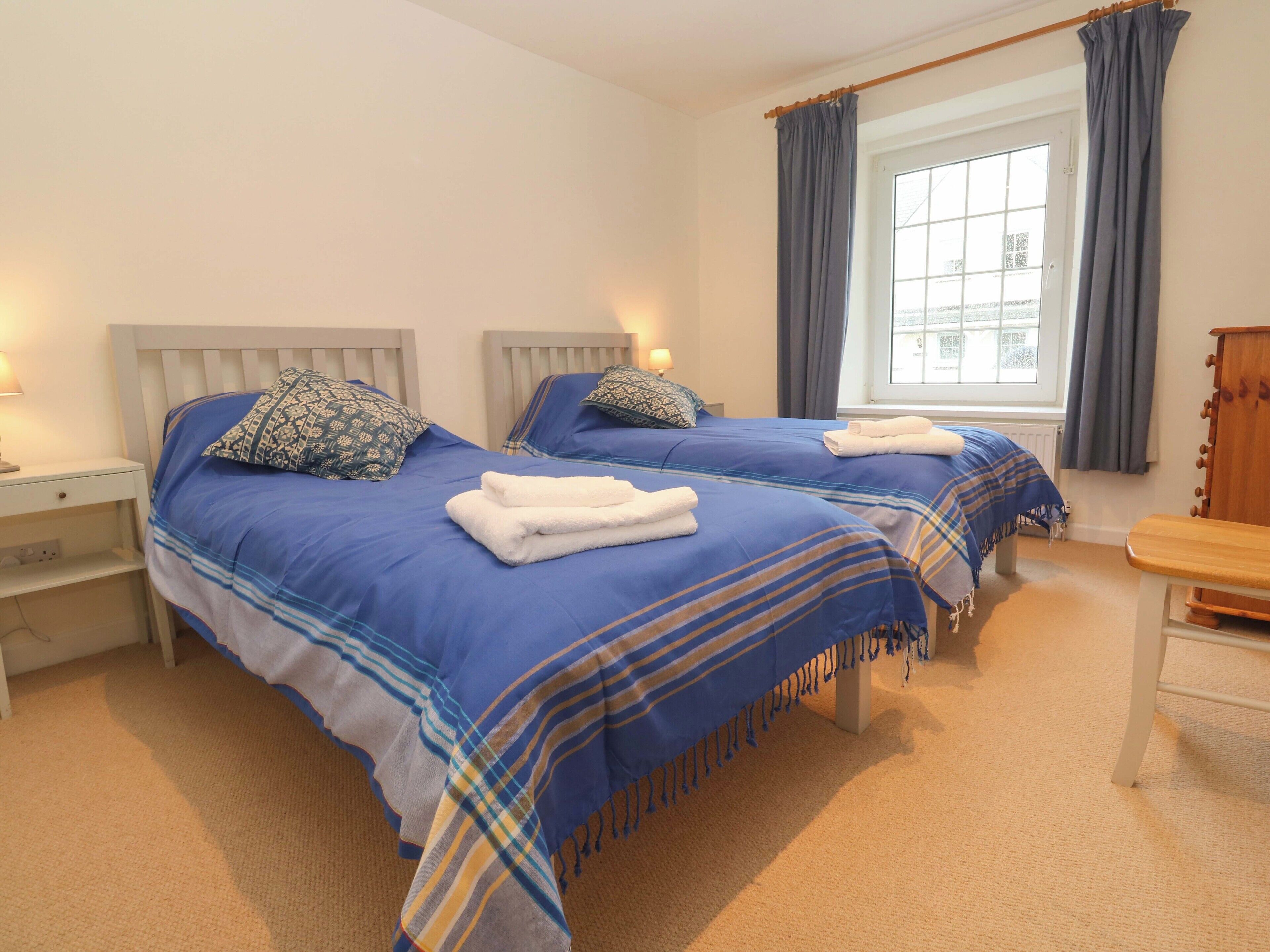 A hotel room with two beds, each with a blue bedspread and matching pillows, a wooden nightstand, a small wooden table, and a window with curtains.