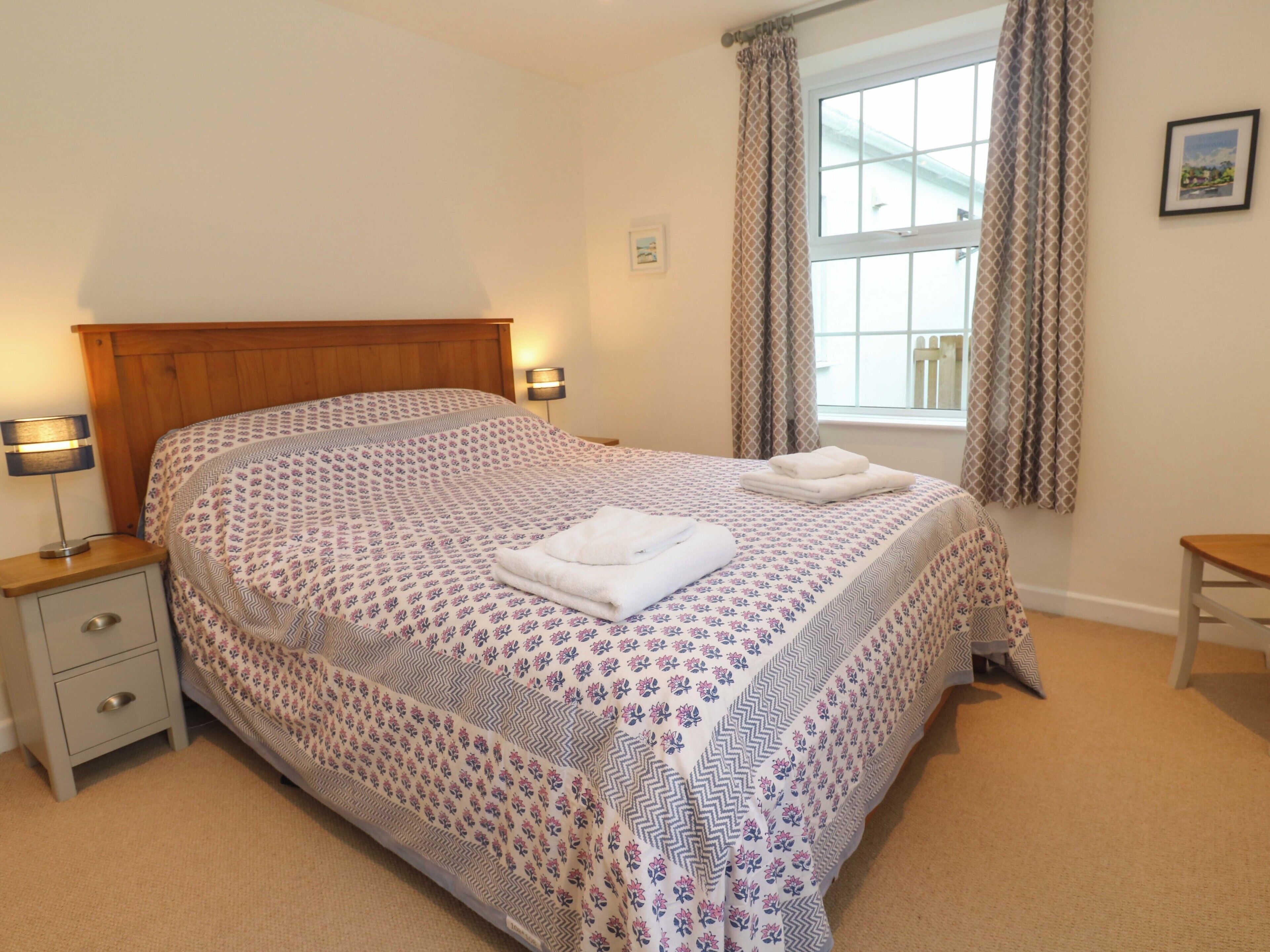 A neatly made bed with a patterned bedspread, two bedside lamps, a nightstand with drawers, a window with curtains, and a framed picture on the wall.