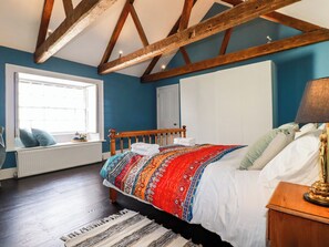 A bedroom with a wooden ceiling, a large window, a bed with a colorful blanket, and a nightstand with a lamp. - Carew House Cottage (Redruth)