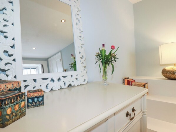 A room with a white vanity, a mirror with ornate white frame, a vase with flowers, and a lamp on a shelf. - Carew House Cottage (Redruth)