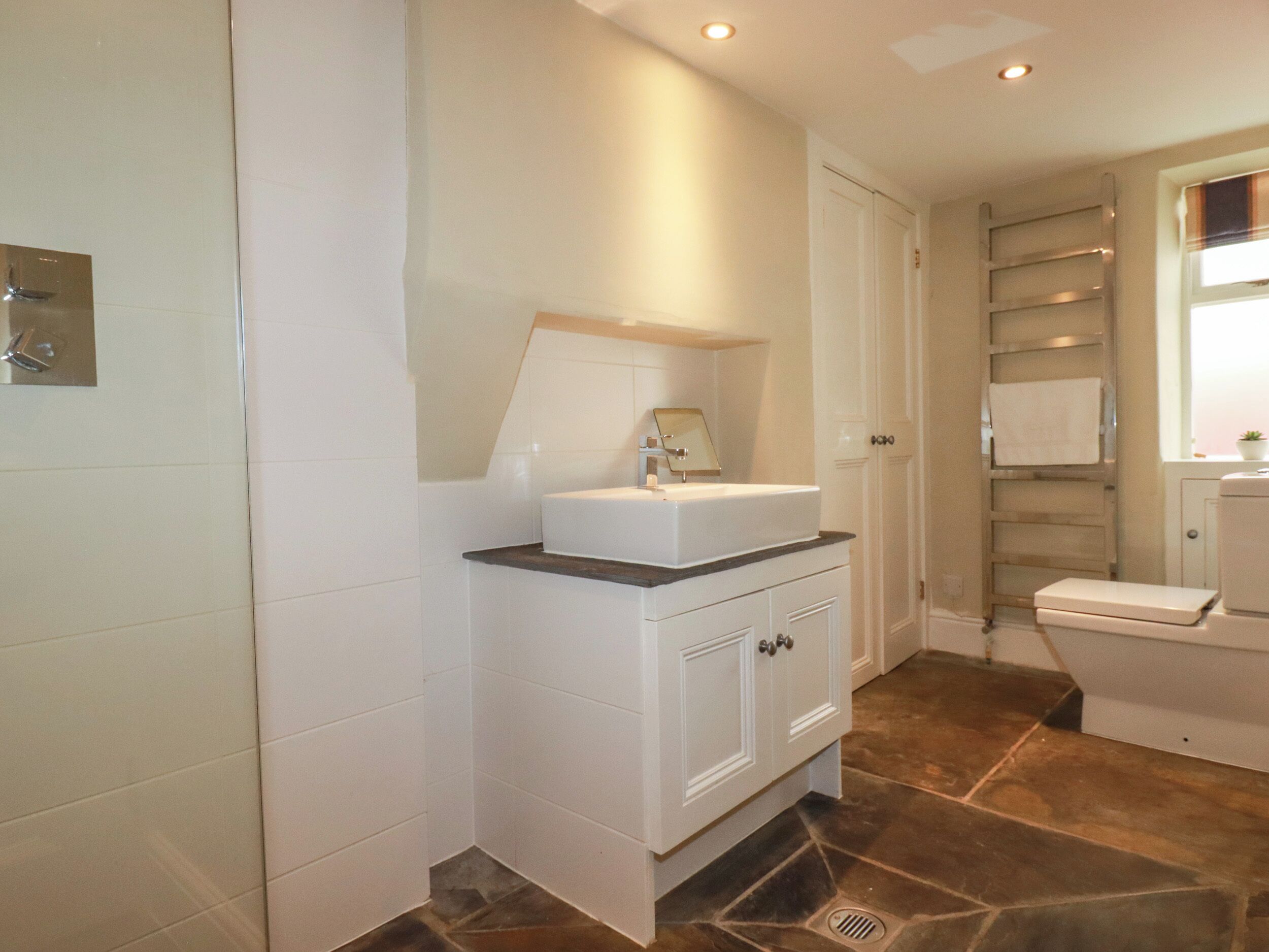 Baño con muebles blancos, un lavabo y un espejo. Se ve un perchero y un inodoro.