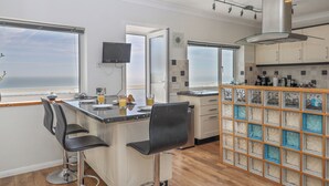 A modern kitchen with a large island, bar stools, a TV, and a view of the ocean. - Seahaze (Torpoint)