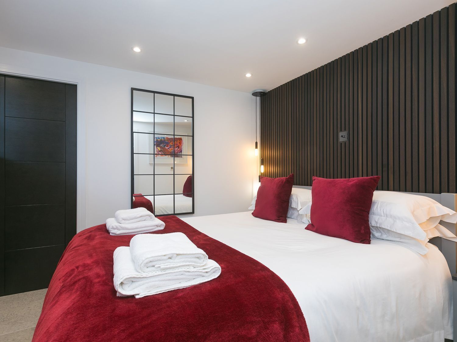 Quarto de hotel moderno com uma cama grande, roupa de cama vermelha e branca, um espelho e um painel de parede de madeira.