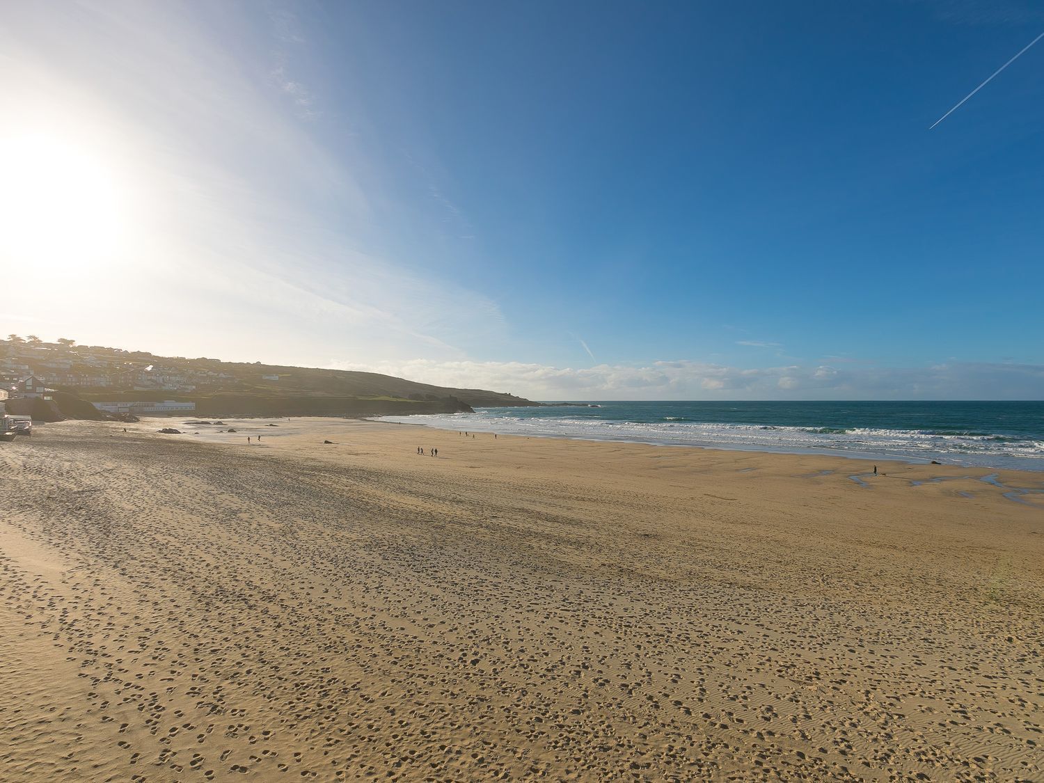 A wide sandy beach with footprints, a clear sky, and a distant shoreline with buildings.