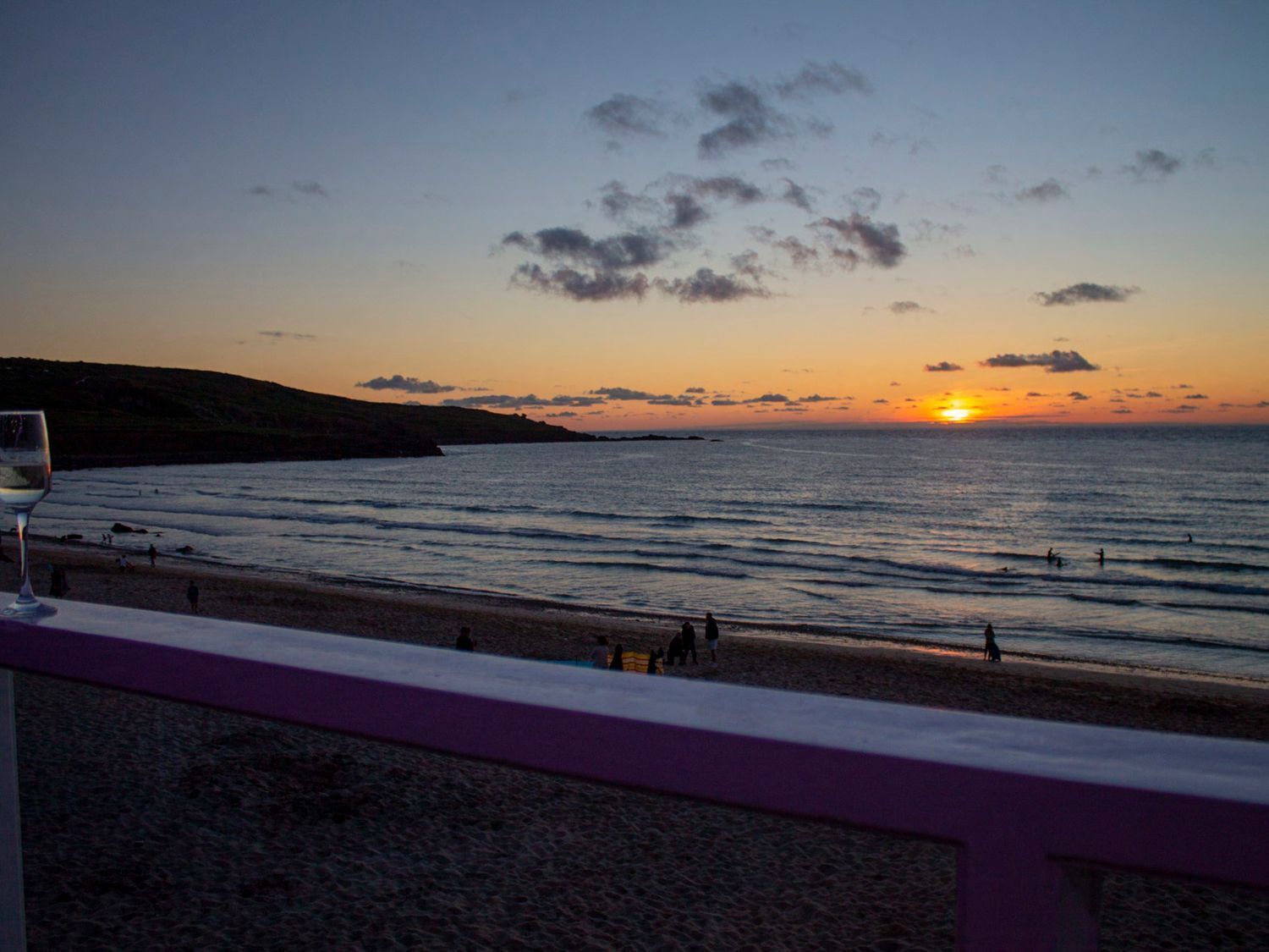 A sunset view from a balcony overlooking a beach with people in the water and a glass of wine on the table.